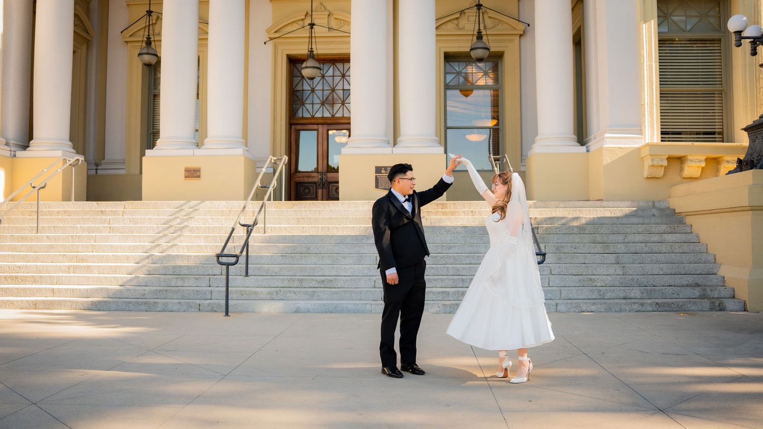 A couple in formal wedding attire dances together on grand stone steps of a historic building with columns.