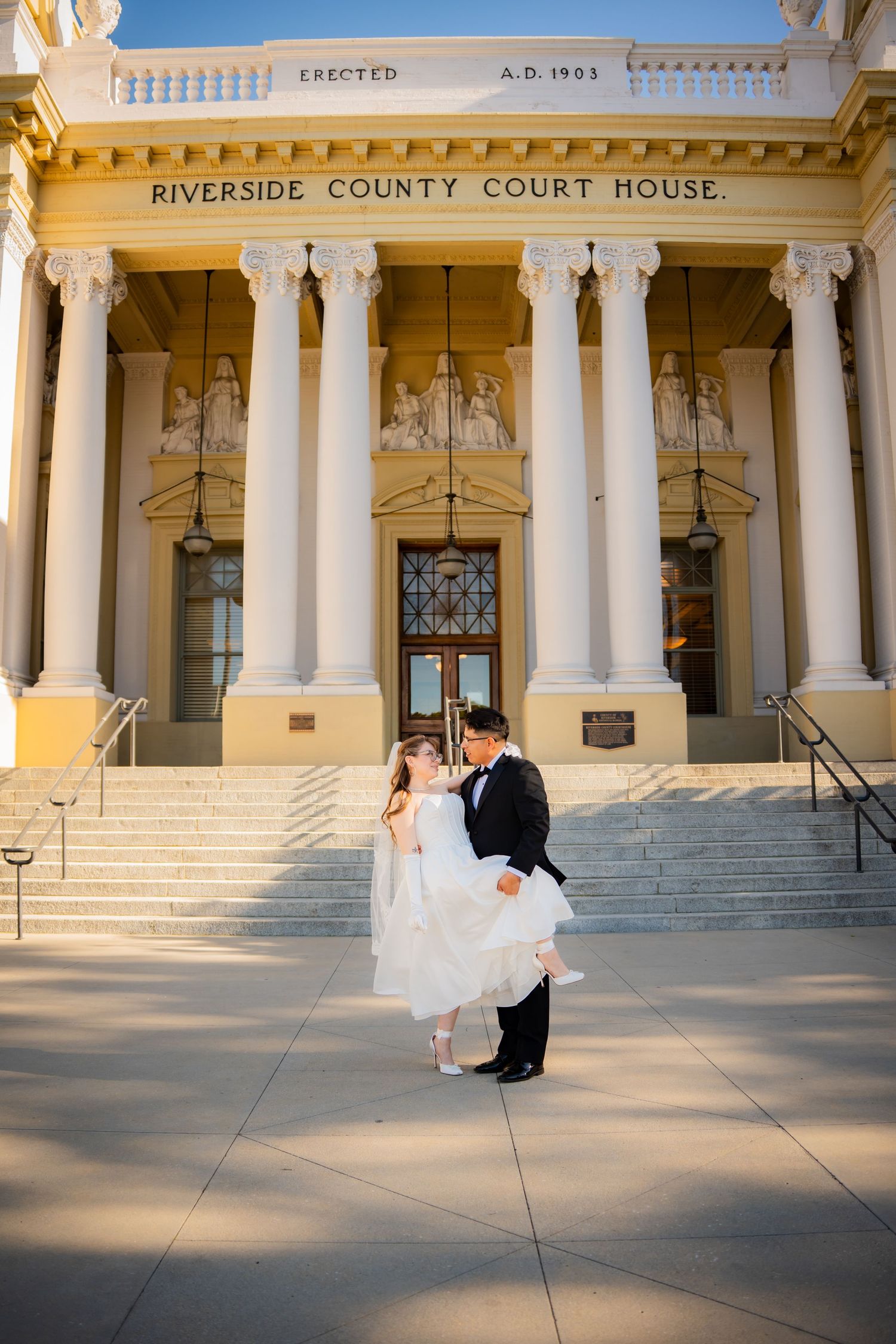 A couple in formal wedding attire embraces on the grand steps of the historic Riverside County Court House.