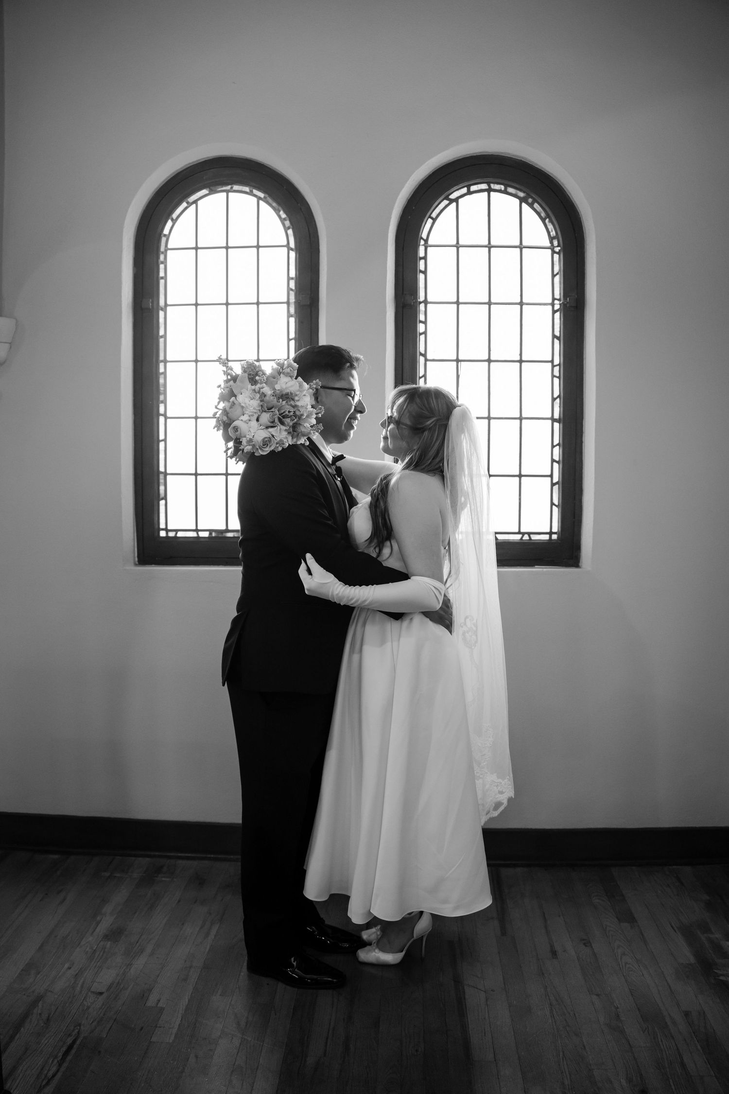 A black and white wedding photo shows a couple in formal attire standing before two arched windows.