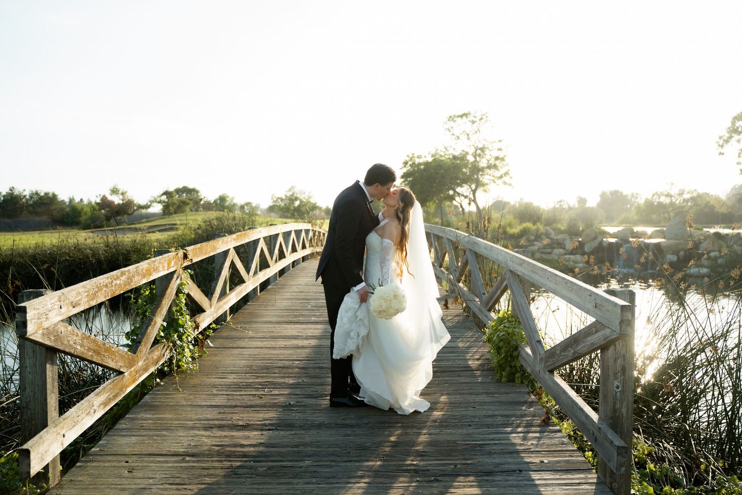 Romantic couple in wedding attire share intimate moments on a wooden bridge at sunset over tranquil waters.
