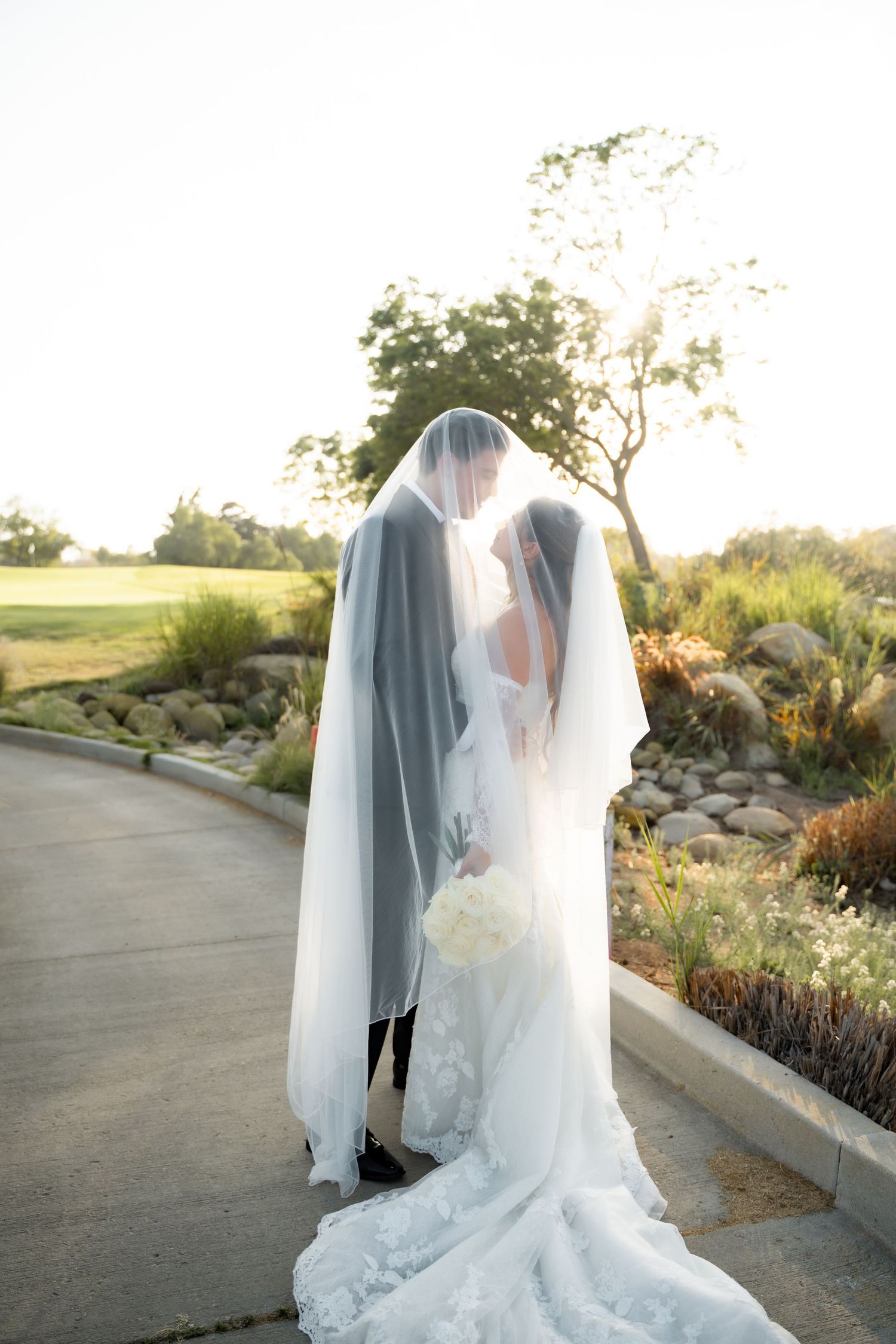 A romantic wedding couple in white attire walk along a winding garden path at sunset.