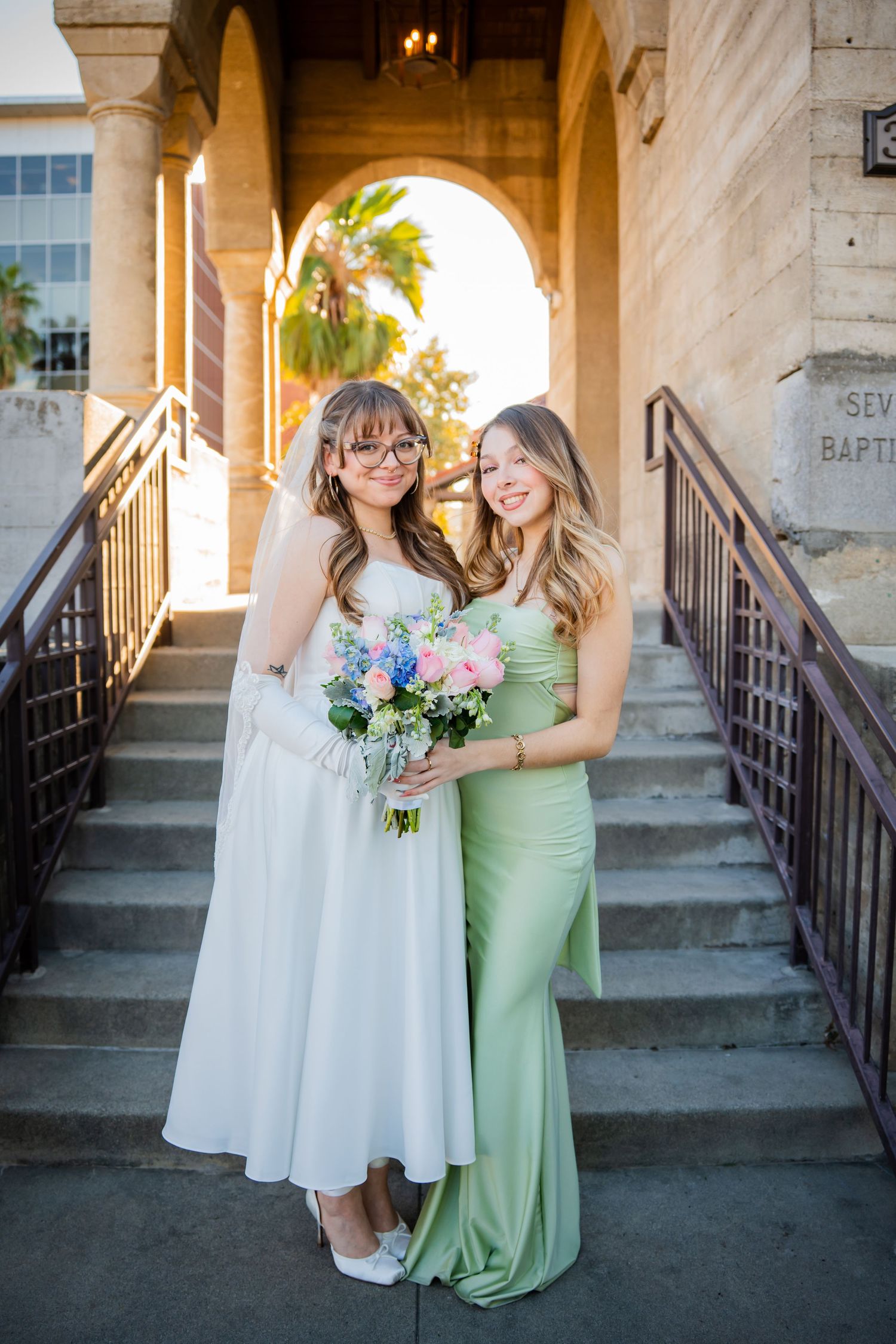 Two people pose on church steps in formal attire, one wearing a white dress and one in mint green, holding a floral bouquet.