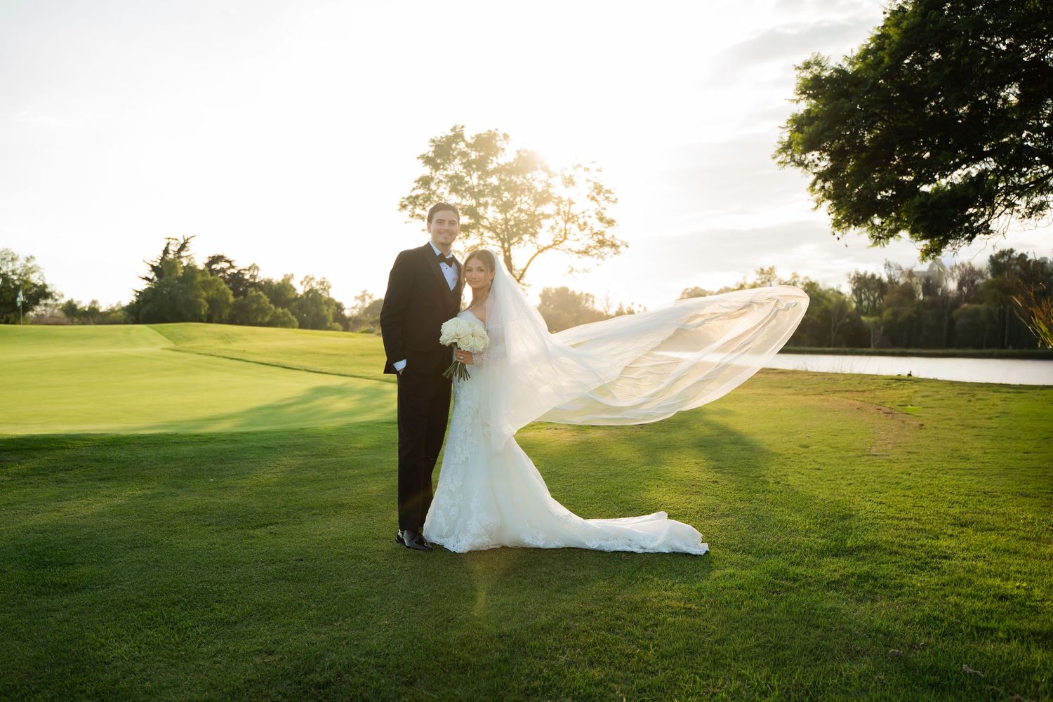 A couple in wedding attire stands silhouetted against the sunset on a lush green golf course.