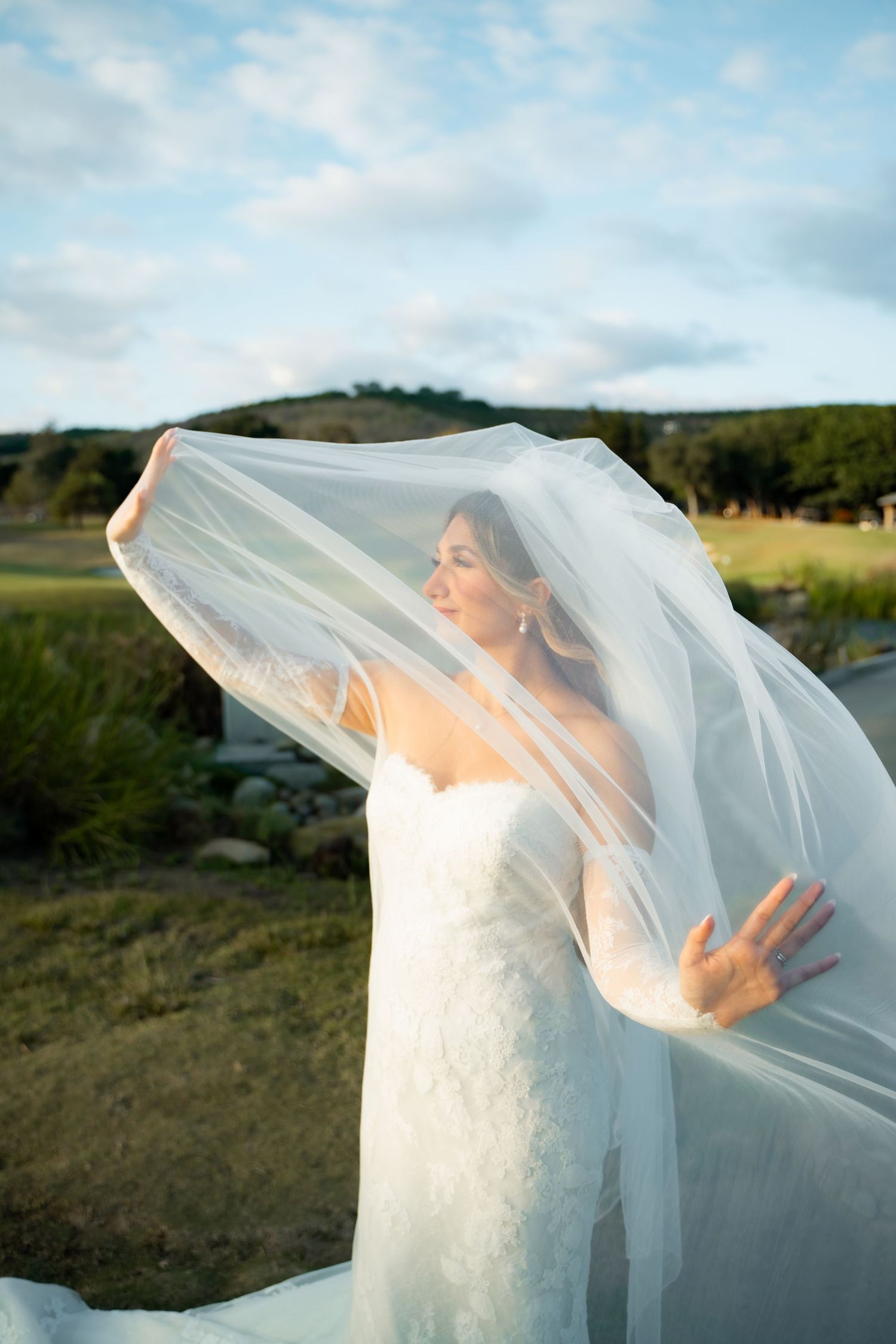 A bride in a white wedding dress plays with her flowing veil against a scenic outdoor landscape at sunset.