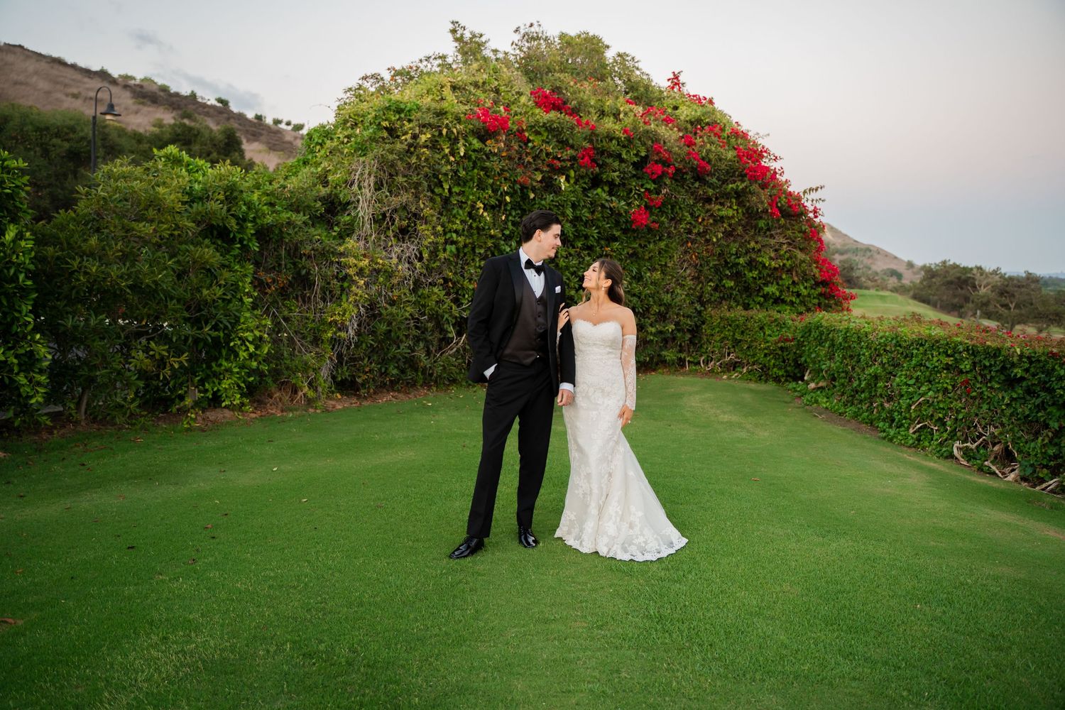 A scenic outdoor wedding ceremony on a lush green golf course with vibrant red flowers blooming in the background.