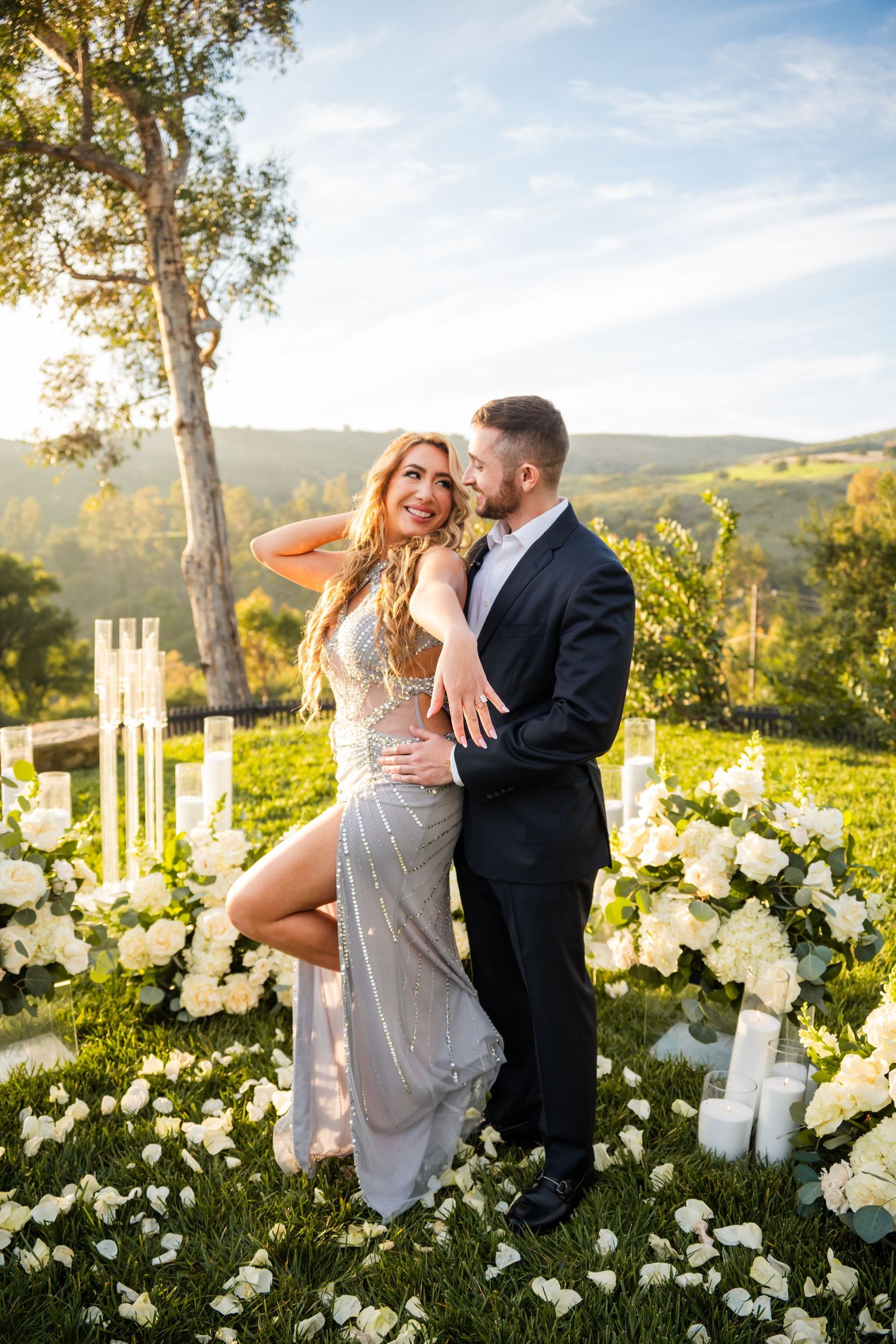 Romantic engagement photoshoot in a garden setting with white roses and mountain views at sunset.