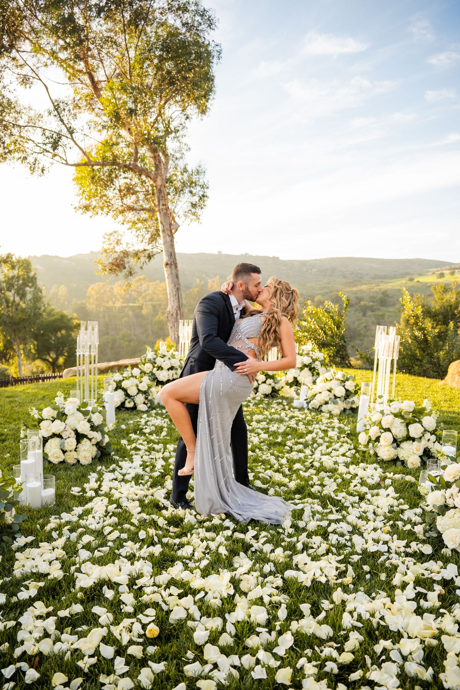 Romantic couple shares a dip kiss among white floral arrangements on a scenic hilltop at sunset.