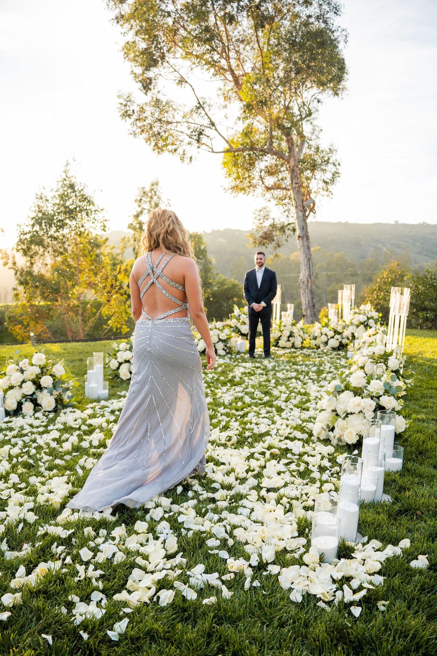 A bride in a lavender dress walks through a field of white flowers during an outdoor wedding ceremony at sunset.