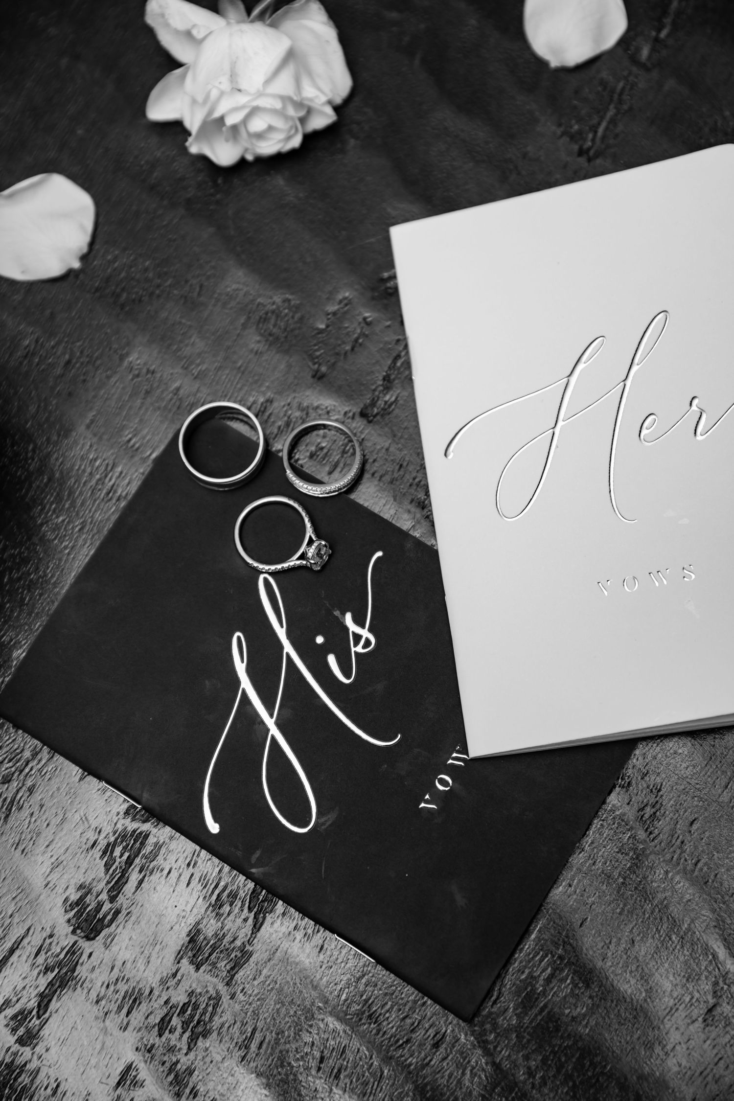 Black and white wedding photo of rings and calligraphy cards reading 'His' and 'Her' with rose petals on dark surface.