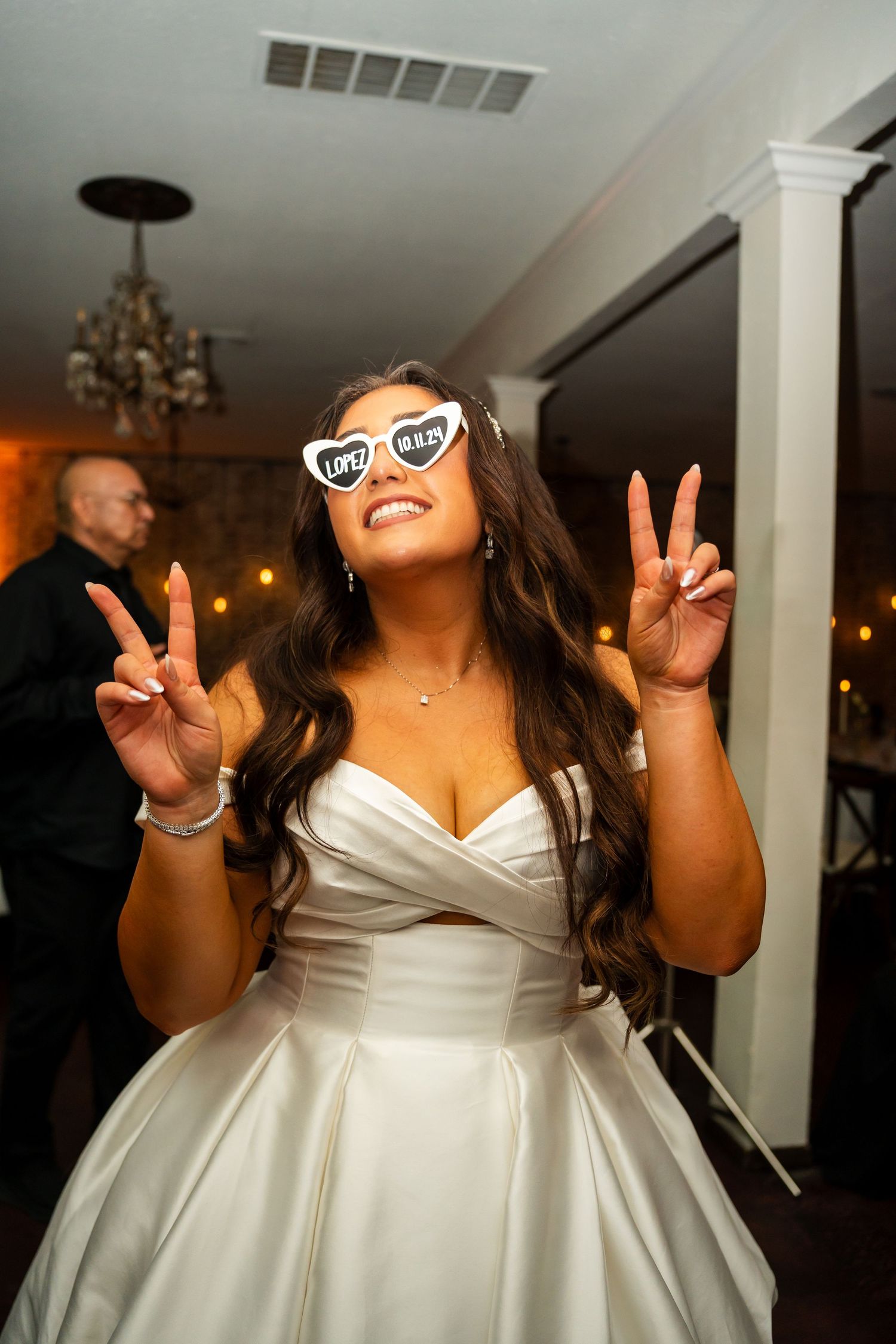 A joyful bride in a white satin gown shows peace signs while laughing and celebrating at her wedding reception.