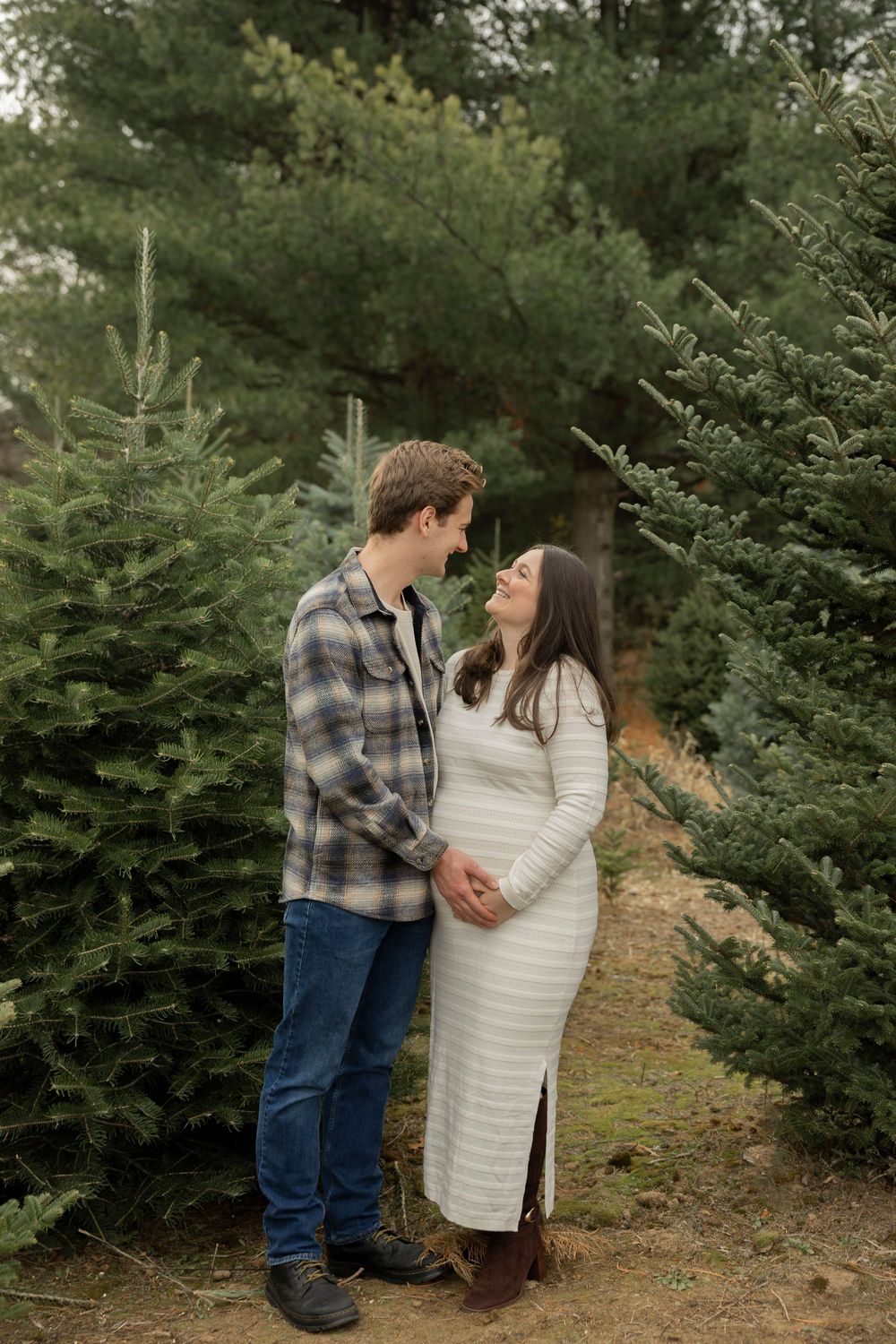 A pregnant couple shares tender moments among evergreen trees at a Christmas tree farm during winter.