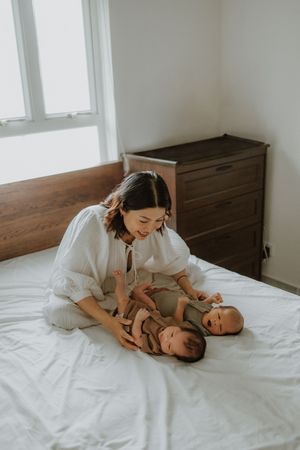 A smiling mother in white sits on a bed with two newborn babies lying in her lap in a bright bedroom.