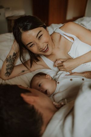 Intimate candid moment shows mother breastfeeding baby while lying down in bed with warm lighting and tattoo visible.
