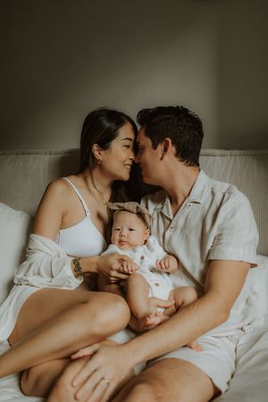 Intimate family moment captured in soft lighting as parents cuddle with their baby on white bedding.