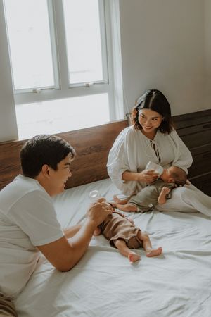 Young couple sitting on a white bed smiling at their newborn baby in a bright, sunlit bedroom.