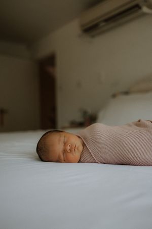 Tiny sleeping newborn baby wrapped in soft pink knit blanket resting peacefully on white bedding.