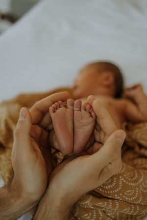 Adult hands gently cradle tiny newborn baby feet against soft beige blanket in tender moment.