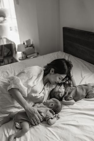 Black and white photo of a mother tenderly kissing her newborn baby while lying on a white bed.
