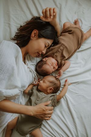 Mother lying on white bed with two sleeping newborn twins in neutral onesies, sharing a tender moment.