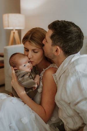Loving parents share tender kisses with their baby while lying together on a bed in warm natural lighting.