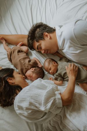 Two parents lying on a white bed, lovingly gazing at their newborn twins in soft neutral outfits.