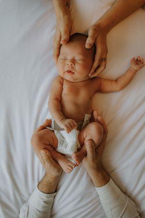 Gentle hands hold and comfort a peaceful sleeping newborn baby wearing a diaper on white bedding.
