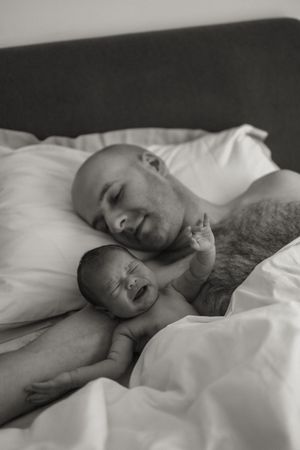 A tender black and white portrait of a parent and crying baby lying together on white bedding.