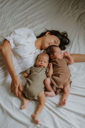 Mother resting on white bed with two sleeping newborn babies in olive and brown ribbed onesies.