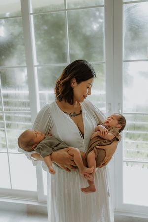 A mother in a white dress holds two newborn babies wrapped in neutral-toned blankets near a bright window.