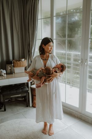 A woman in a white dress holds two newborn babies near a bright window in a serene, modern room.