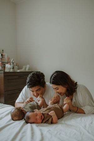 A couple lies on a white bed smiling at their newborn baby, sharing a tender family moment in a cozy bedroom.