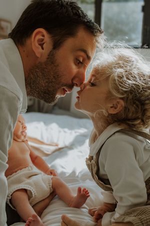 A tender family moment as parent and child share an intimate nose-to-nose moment while a baby rests nearby on white bedding.