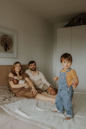 Family relaxing together on bed with young child in denim overalls standing while others sit in casual home setting.