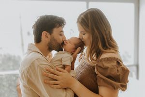 Parents share tender moment with their newborn baby beside bright window in warm natural lighting.