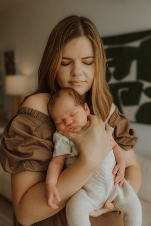 Parent in brown top tenderly embraces sleeping infant wearing white onesie in intimate indoor portrait series.