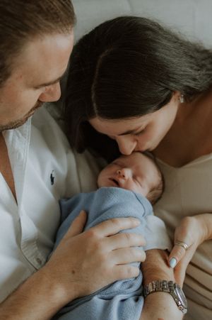 New parents tenderly kissing their sleeping newborn baby wrapped in a soft blue swaddle blanket.