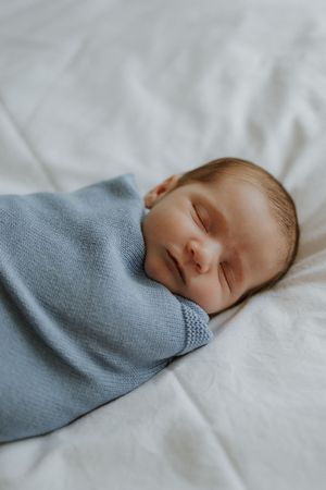 Peaceful newborn baby sleeping soundly, swaddled in a soft blue knit blanket on white bedding.