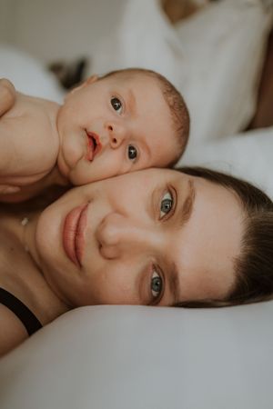A tender close-up portrait shows an adult and baby lying together on white bedding in soft, warm lighting.