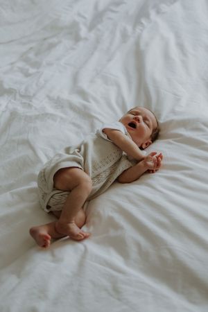 A yawning newborn baby lying on white bedding wearing a neutral linen romper with bare feet.