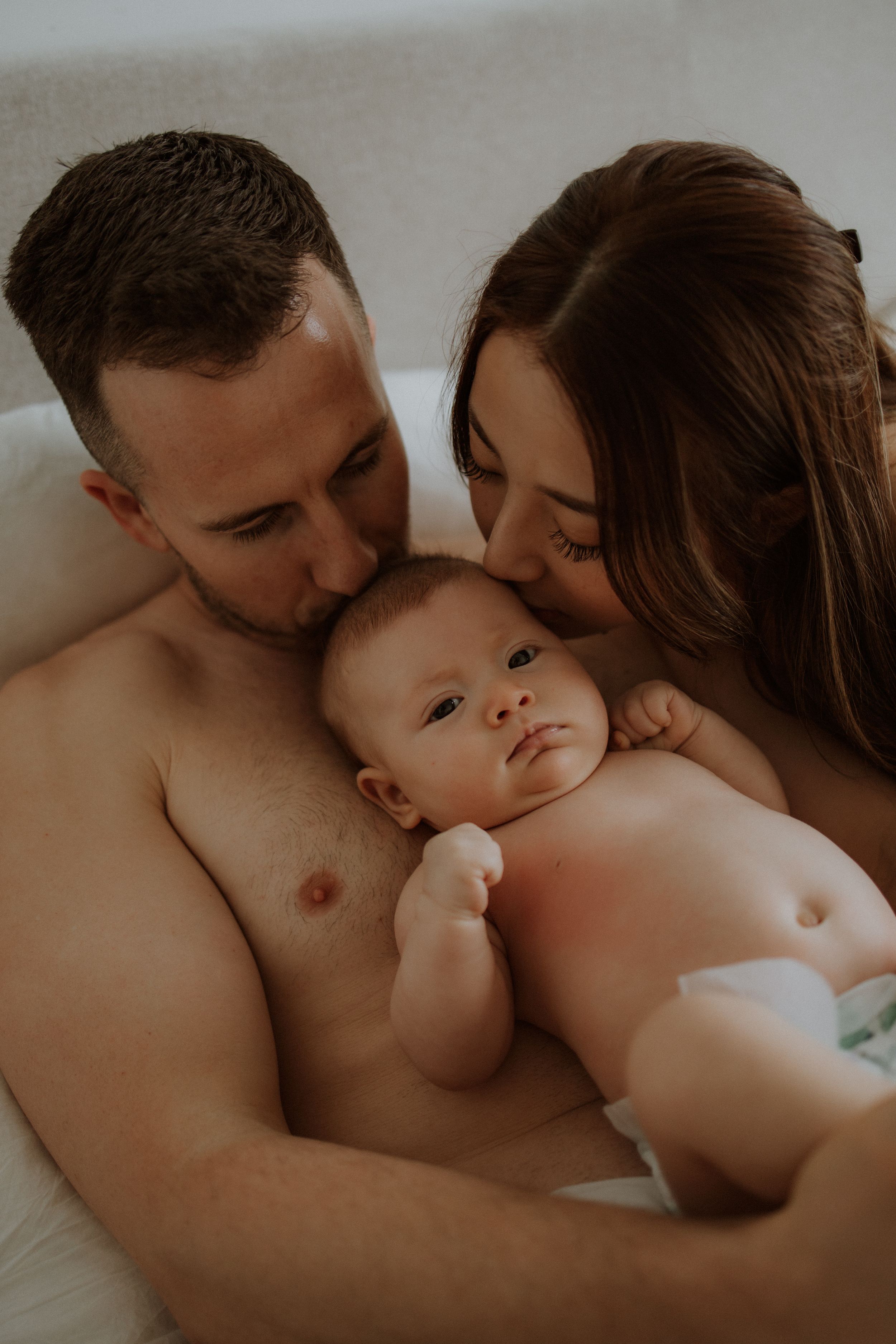 Intimate family portrait showing tender moment between parents and their small baby in soft lighting.