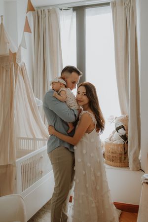 A couple shares an intimate moment near light-filled windows in a bright nursery with soft beige curtains and decorative bunting.