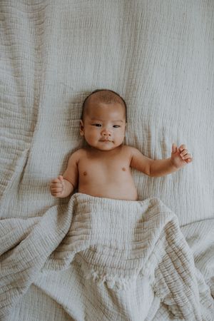 Adorable baby lying on a soft white muslin blanket, looking at the camera with one hand raised.