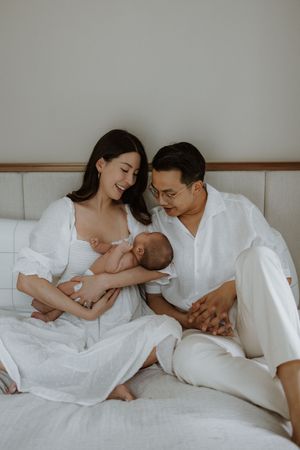 A couple in white outfits sit on a bed, lovingly gazing at their newborn baby in a serene nursery setting.