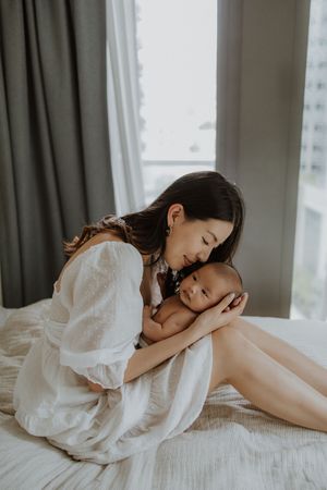 A mother in a white dress tenderly holds her newborn baby while sitting on a bed near a window.
