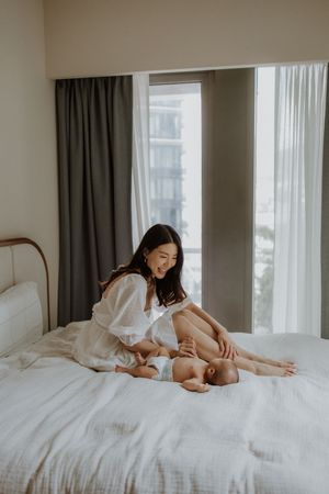 A smiling Asian mother in white dress sits on bed playing with her newborn baby in a bright modern bedroom.