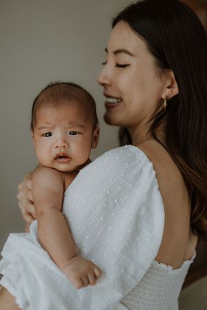 A smiling Asian mother in a white dress tenderly holds her newborn baby against her shoulder.