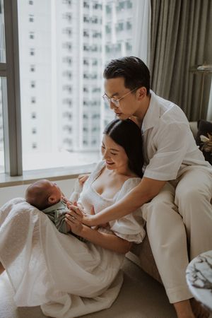 A couple sitting by a window, lovingly gazing at their newborn baby in a bright, modern apartment.