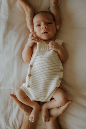 A baby wearing a white knit romper lies on white bedding while being gently held by adult hands.