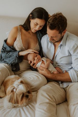 A couple in casual attire cuddles with their newborn baby on a cozy bed while their dog rests beside them.