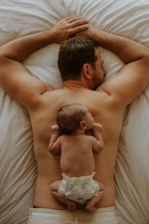 Newborn baby in diaper lies on father's back while both rest peacefully on white bedding.